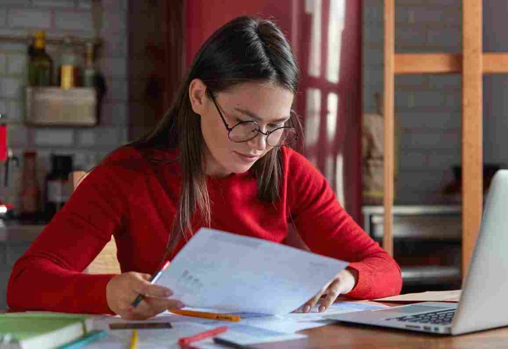 horizontal shot project manager spectacles red juumper looks attentively documents thinks how attract clients increase income poses against kitchen interior with portable laptop 11zon