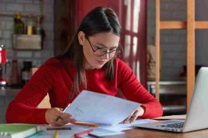 horizontal shot project manager spectacles red juumper looks attentively documents thinks how attract clients increase income poses against kitchen interior with portable laptop 11zon